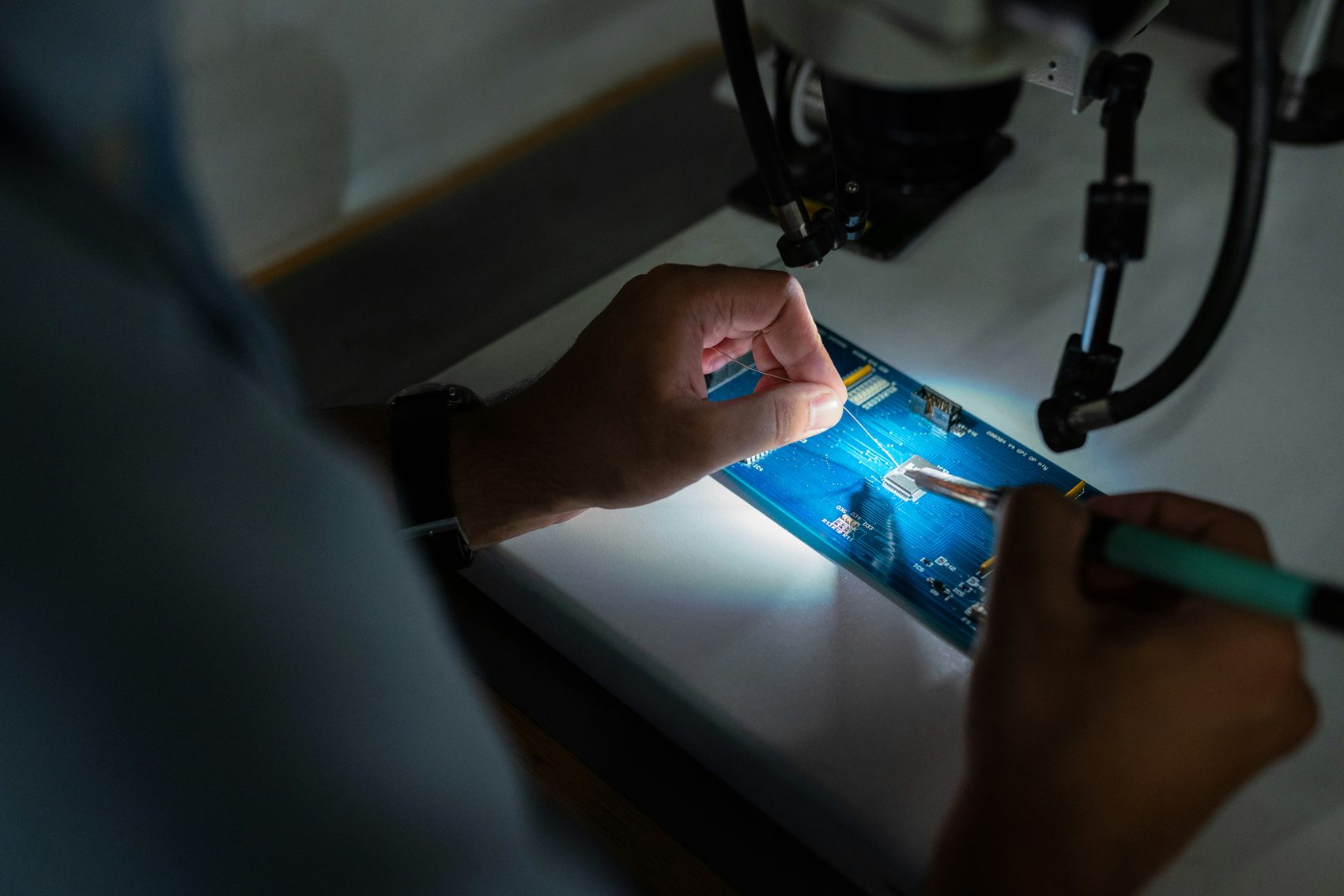 Two hands soldering a fine wire onto a lit blue circuit board under a bench microscope