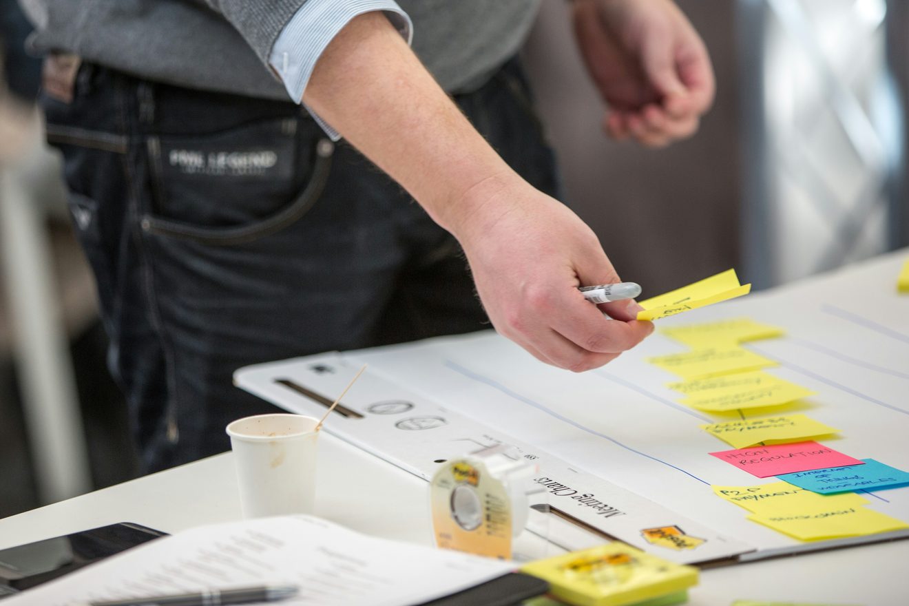 A hand placing a yellow sticky note onto a whiteboard already covered in handwritten notes