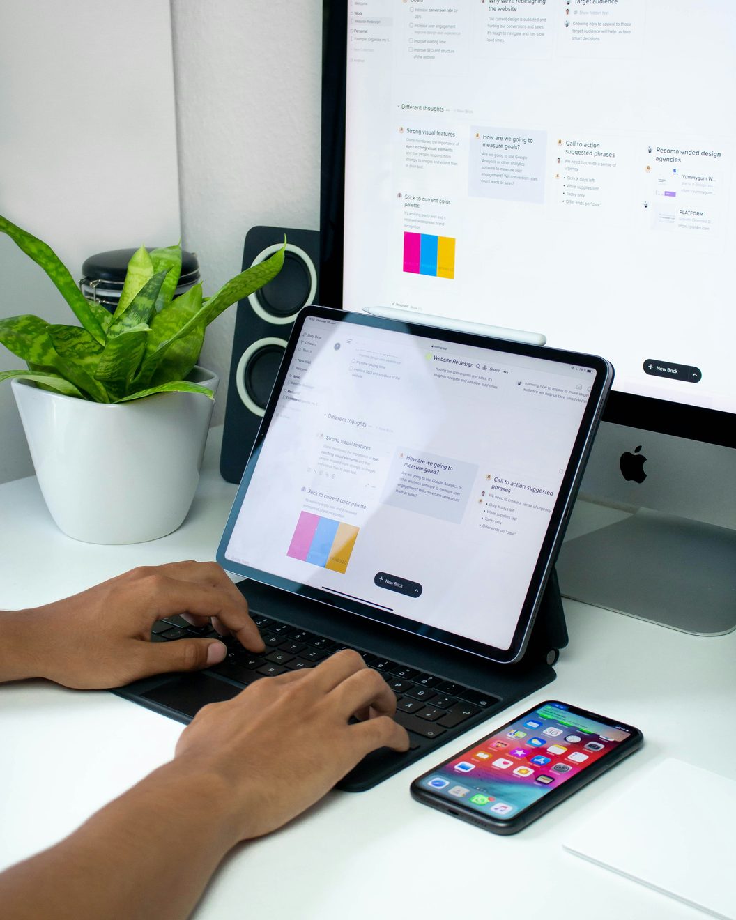 Hands typing on a tablet keyboard beside a monitor showing a project board and a potted plant