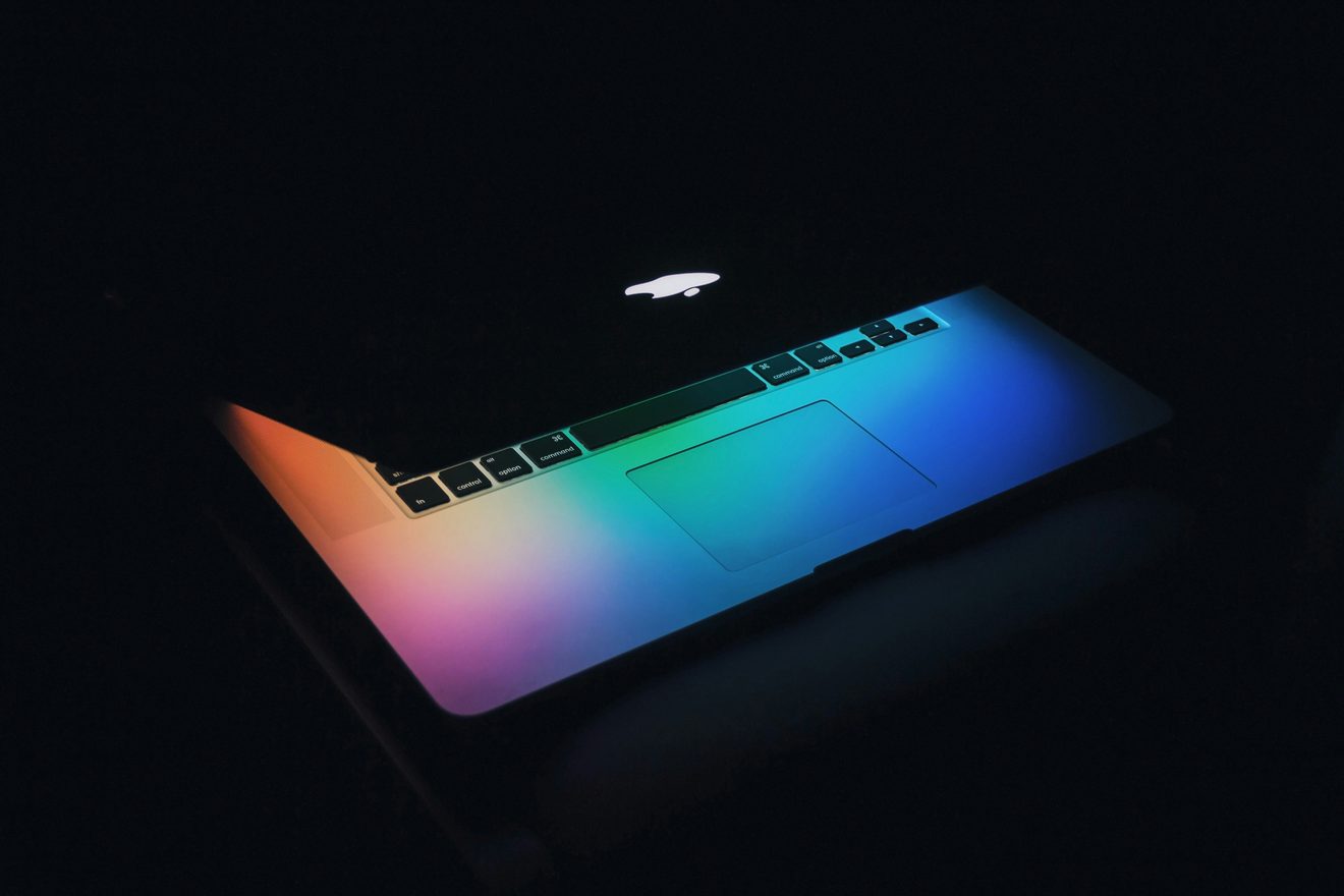 An open laptop in a darkened room lit only by a rainbow gradient glowing from its screen