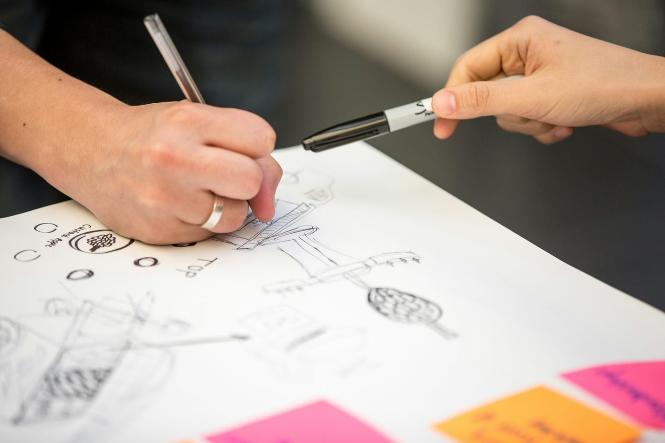 Two people sketching product concepts in marker on a large sheet of paper, one passing a pen across