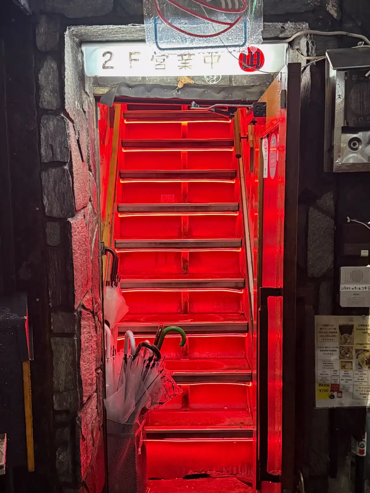 Steep stairs lit up bright red, with a sign saying the second-floor bar is open and wet umbrellas in a bin by the door.