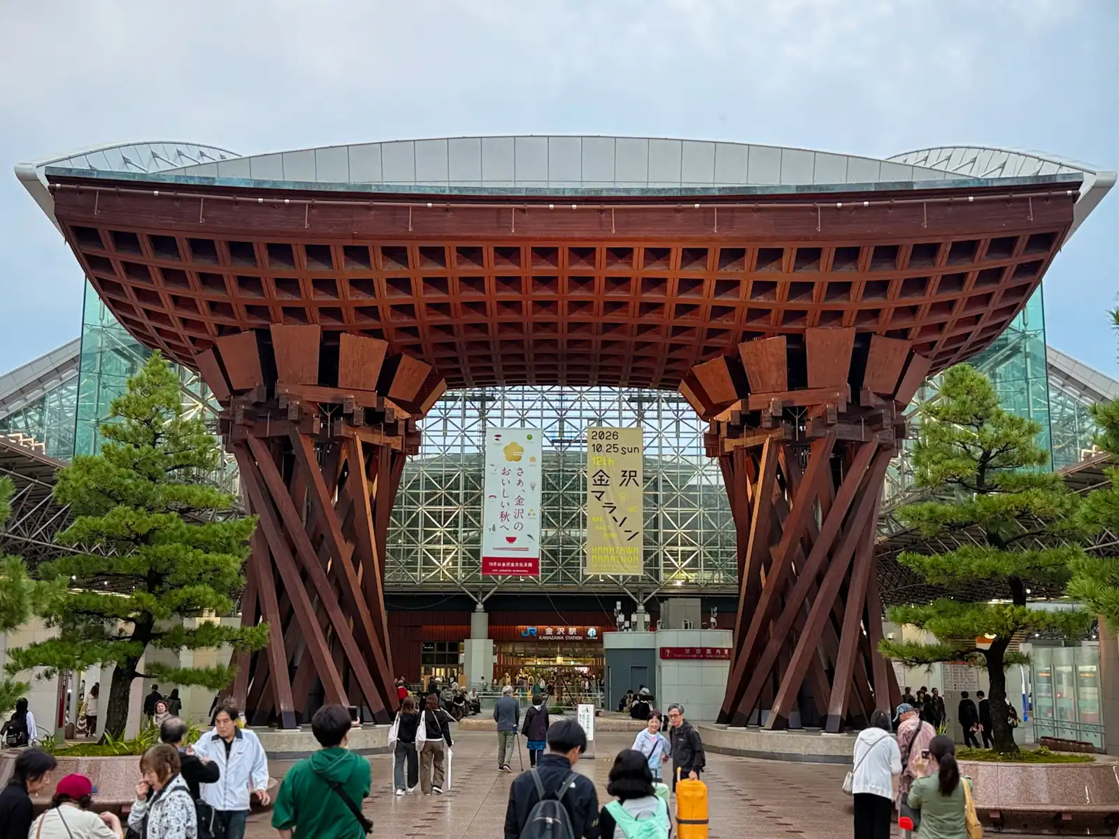 The Tsuzumi Gate from the front, with pine trees on both sides and the Motenashi Dome behind it. People walk to and from the station entrance under the gate.