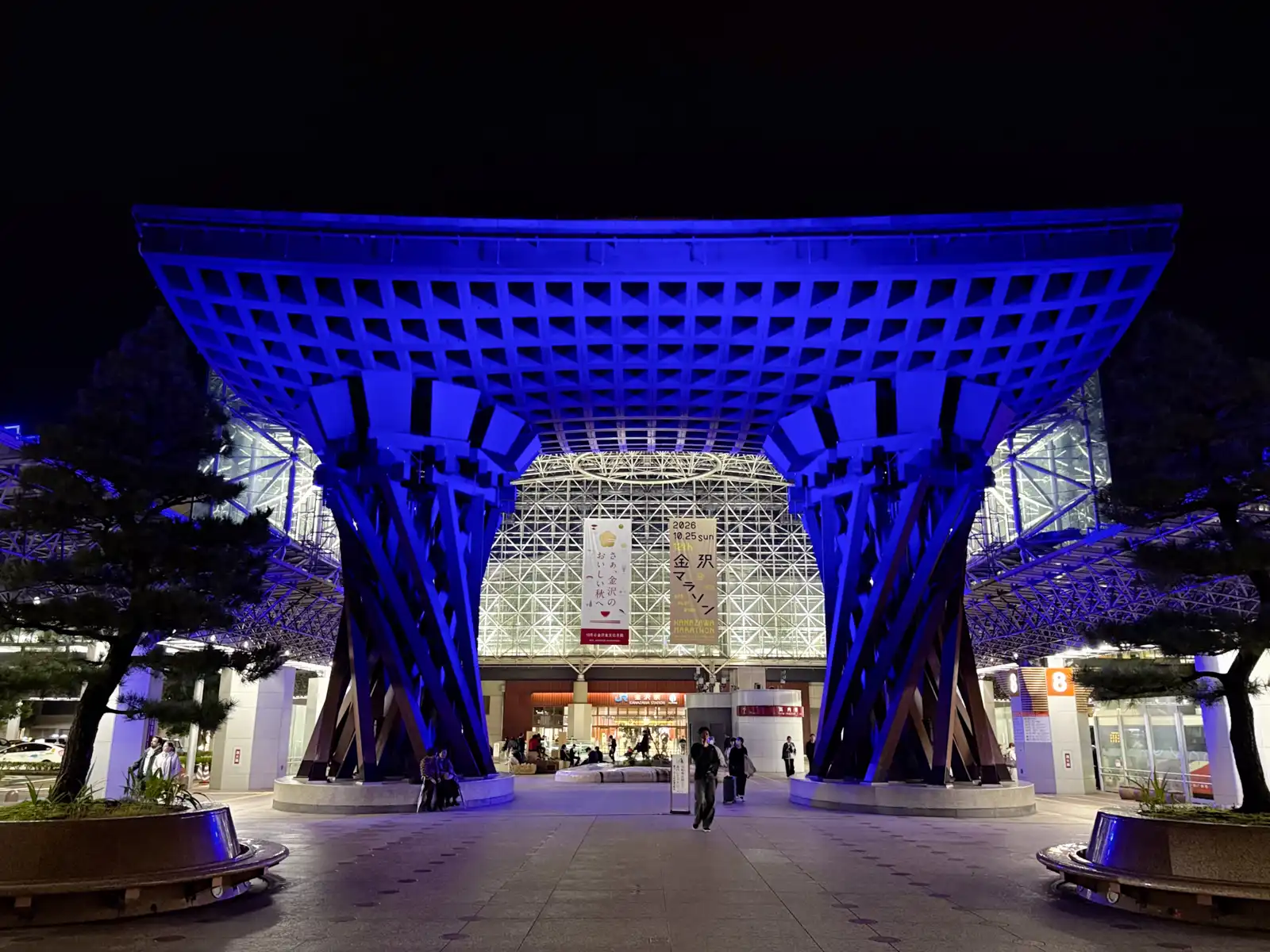 The Tsuzumi Gate lit in blue at night, with the Motenashi Dome glowing behind it and pine trees on both sides.